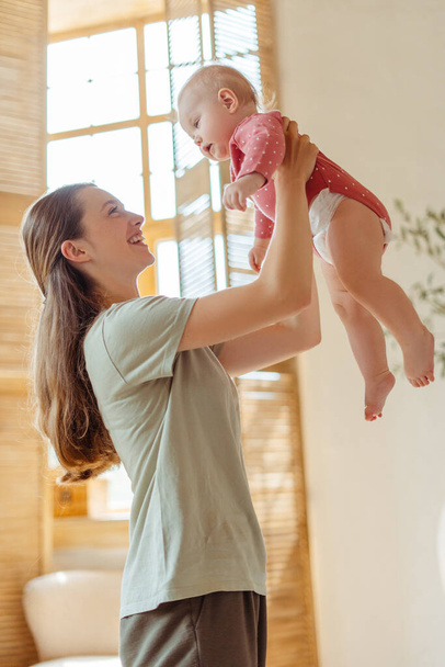 Hermosa madre joven sonriente sosteniendo, jugando con su pequeño bebé lindo en casa, divirtiéndose. Amor, cuidado, concepto de paternidad - Foto, Imagen