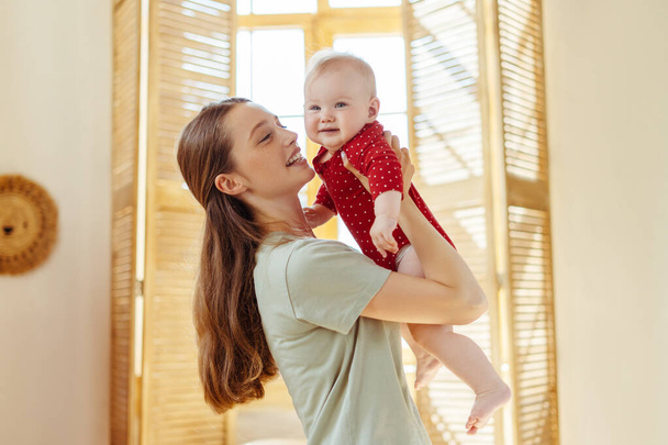 Feliz, sonriente madre sosteniendo a su lindo bebé en la sala de estar en casa. Niñera joven positiva cuidando a un bebé hermoso. Desarrollo infantil, cuidado, amor - Foto, Imagen