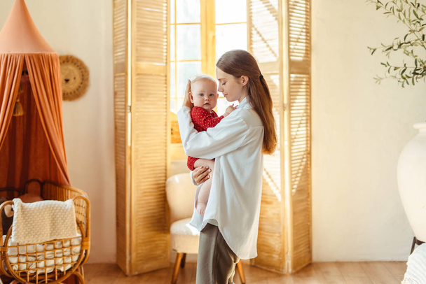 Bela jovem mãe com olhos fechados segurando pequeno bebê na sala de estar em casa. Mulher atraente cuidando da criança. Conceito de amor, família, paternidade - Foto, Imagem