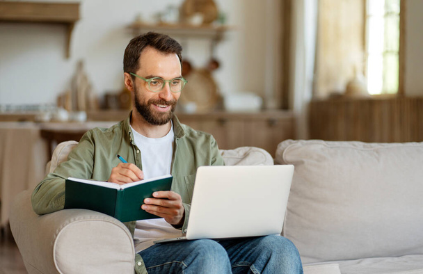 Handsome smiling bearded businessman wearing eyeglasses using laptop computer, taking notes, watching training courses at home. Smart student studying, learning language, online education concept - Photo, Image