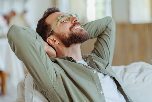 Millennial caucasian handsome bearded man, wearing trendy glasses, sitting on sofa and stretching hands at workplace or loft apartment. Professional occupation, freelance  - Photo, Image