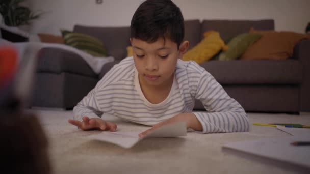 Menino latino concentrando-se em fazer origami deitado no chão em casa. Criança no acolhedor tapete da sala de estar fazendo avião de papel desfrutando de seu tempo livre. Conceito de hobbies infantis positivos em casa. - Filmagem, Vídeo