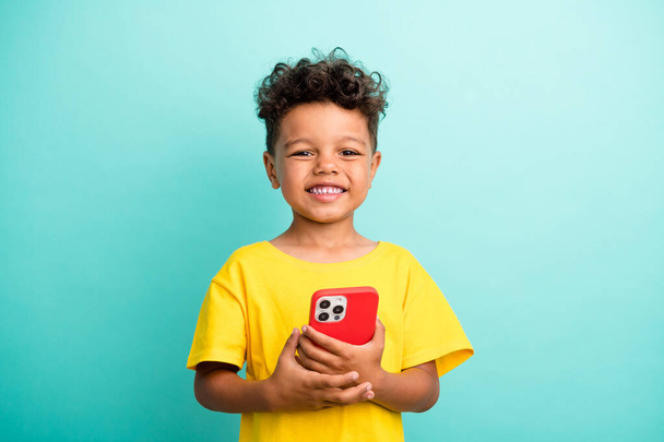 Portrait of satisfied small boy with curly hairstyle wear stylish t-shirt holding smartphone isolated on turquoise color background. - Foto, Imagem