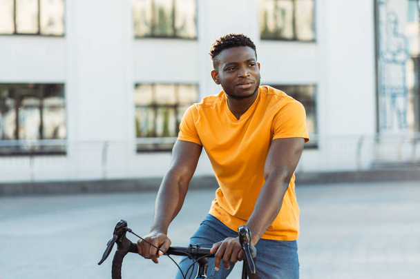 Портрет красивого усміхненого афроамериканця в помаранчевій сорочці, що їде на велосипеді на міській вулиці, дивлячись подалі. Привабливий здоровий спосіб життя чоловіків. Концепція спорту, хобі, транспорту - Фото, зображення