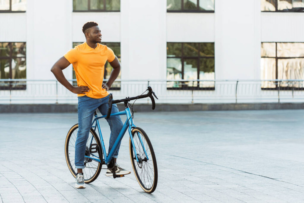 Σκούρο δέρμα όμορφος άντρας περπατά στο δρόμο κοντά στο ποδήλατο ενώ κοιτάζει αλλού. Ηρεμία αφροαμερικανός φοιτητής διασταύρωση με ποδήλατο  - Φωτογραφία, εικόνα