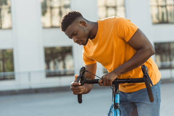 Portret i jakiś Afrykanin sprawdzający kierownicę roweru, stojący na miejskiej ulicy. Atrakcyjny mężczyzna w stylowych ubraniach, ulubione hobby. Koncepcja jazdy na rowerze, naprawy rowerów - Zdjęcie, obraz