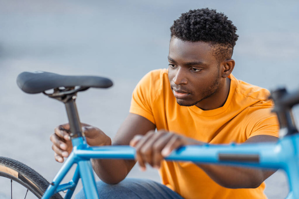 Closeup portrait of attractive Nigerian man checking bicycle seat on street. Young serious male wears orange t-shirt. Concept of cycling - Photo, Image