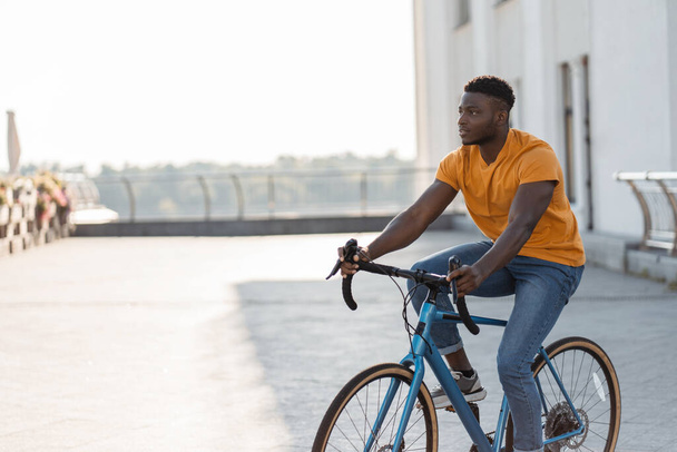 Вид в полный рост на красивого улыбающегося афроамериканца, который едет на велосипеде по городской улице и смотрит в сторону. Спорт, хобби, концепция активного образа жизни  - Фото, изображение