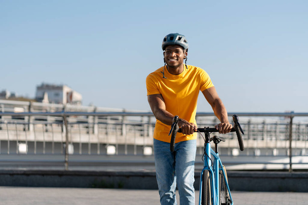 Kentin açık sokaklarında koruyucu kask takan gülümseyen yakışıklı Afrikalı binicinin portresi bisiklet sürüyor. Açık havada şık turuncu tişörtler giyen çekici genç bir öğrenci. Sağlıklı yaşam tarzı kavramı - Fotoğraf, Görsel