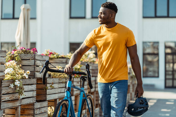 Pozytywny uśmiechnięty Afrykanin trzymający kask ochronny spacerujący z rowerem po ulicy. Atrakcyjny młody student w stylowej, luźnej pomarańczowej koszuli odwracając wzrok, na świeżym powietrzu. Koncepcja rowerowa - Zdjęcie, obraz