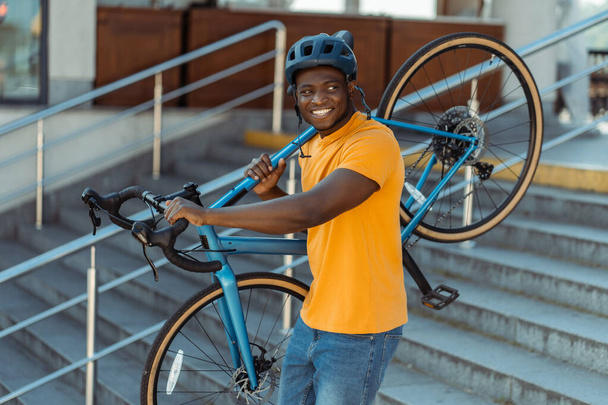 Egy jóképű, mosolygós afro-amerikai férfi portréja, aki sisakot visel, miközben a biciklijét tartja, miközben a városi utcán sétál. Sport, hobbi, aktív életmód koncepció - Fotó, kép