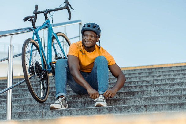 Feliz hombre multirracial con casco protector sentado en las escaleras y mirando hacia otro lado mientras se relaja después de montar en bicicleta - Foto, Imagen