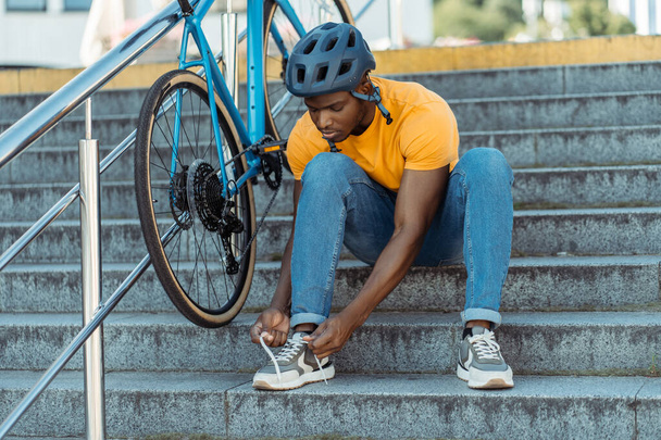 Ernsthafter afrikanisch-amerikanischer Mann, der Schnürsenkel an seine Turnschuhe bindet, während er nach einer Fahrradtour an einer Straßentreppe sitzt. Verkehrs- und Personenkonzept - Foto, Bild