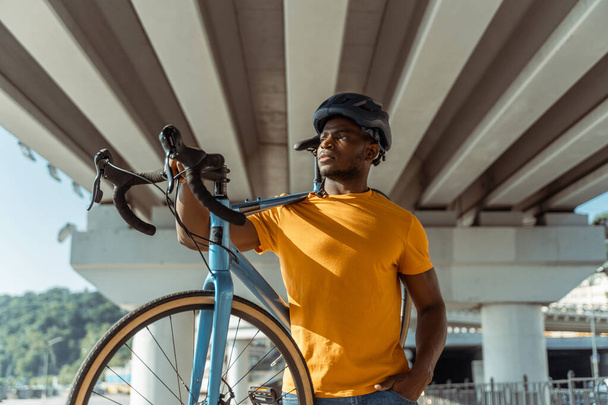 Portret van een knappe lachende Afro-Amerikaanse man met een helm die zijn fiets vasthoudt terwijl hij op straat loopt en wegkijkt. Sport, hobby, actief lifestyle concept  - Foto, afbeelding