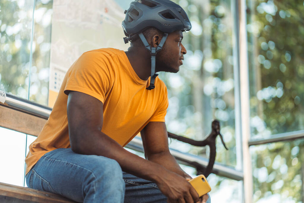 Onnellinen afrikkalainen amerikkalainen mies katselee poispäin ja pitää matkapuhelinta rentoutuessaan linja-autoasemalla polkupyöräilyn jälkeen. Ihmisten elämäntapa käsite - Valokuva, kuva