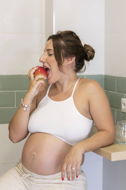 Bella donna incinta vestita con vestiti comodi mangiare una mela rossa in cucina. Foto di alta qualità - Foto, immagini