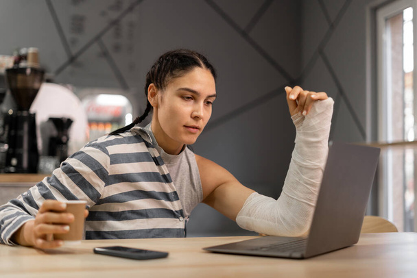 Lächelnde Frau mit gebrochener Hand surft am Laptop im Netz, während sie in einer Bar Kaffee trinkt. Kopierraum - Foto, Bild