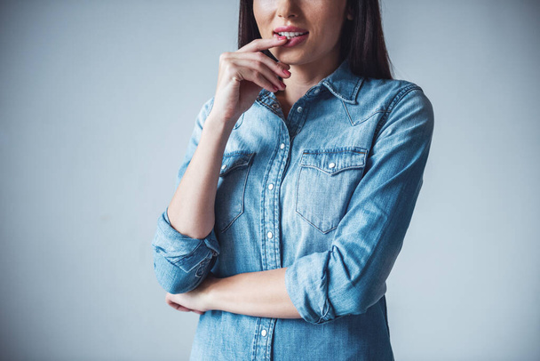Обрезанное изображение красивой девушки в джинсовой одежде, касающейся губ, на сером фоне
 - Фото, изображение