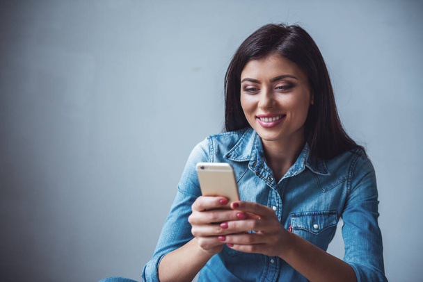 Beautiful girl in jean clothes is using a smartphone and smiling, on gray background - Photo, Image