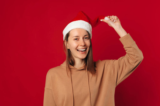 Studio zdjęcie zabawnej kobiety w świątecznym kapeluszu uśmiechniętej do kamery na czerwonym tle. - Zdjęcie, obraz