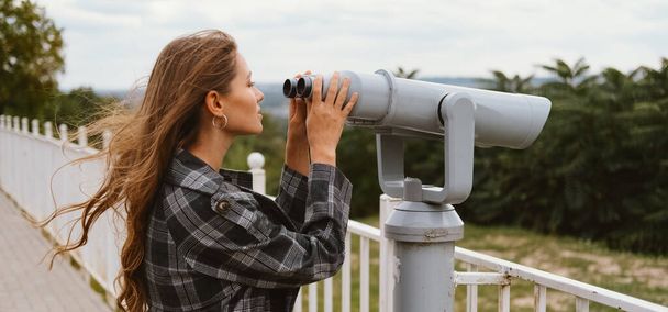 Banner velikost záběr mladé zvědavé ženy při pohledu přes městské dalekohledy na větrný den. - Fotografie, Obrázek
