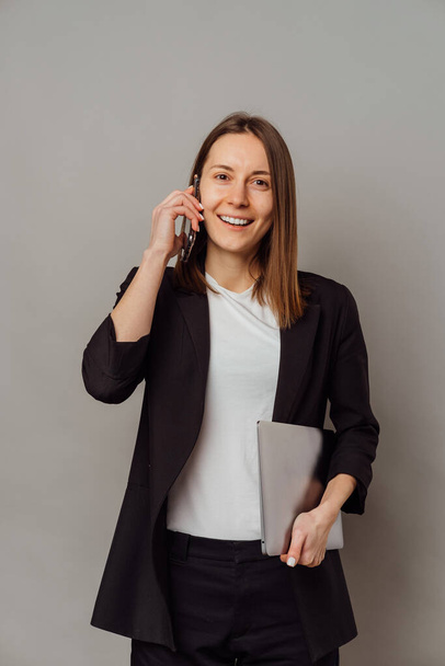 Αυτοπεποίθηση νεαρή χαμογελαστή γυναίκα έχει μια συζήτηση από το τηλέφωνο, ενώ κρατώντας φορητό υπολογιστή πάνω από γκρι φόντο. - Φωτογραφία, εικόνα