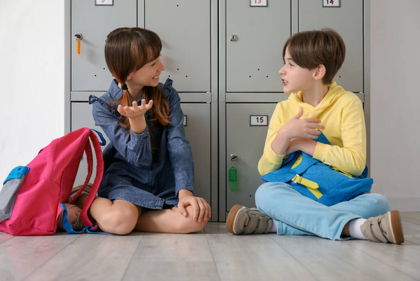Маленькие ученики с рюкзаками сидят возле шкафчика в школе - Фото, изображение