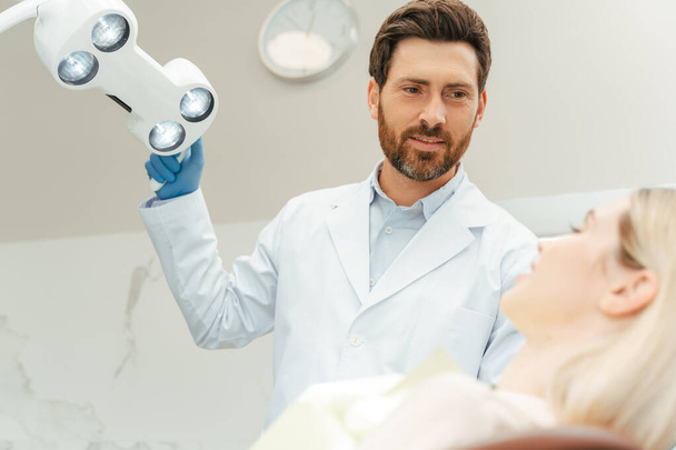 Profesjonalny brodaty dentysta włącza lampę podczas przygotowywania zębów kobiety. Młoda, piękna, uśmiechnięta pacjentka siedząca w nowoczesnej klinice dentystycznej. Leczenie zębów, koncepcja opieki zdrowotnej  - Zdjęcie, obraz