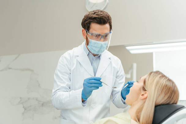 Professional dentist in face mask holding equipment and checking quality of teeth. Young beautiful woman, patient with open mouth sitting in modern dental clinic. Teeth treatment, health care concept  - Photo, Image