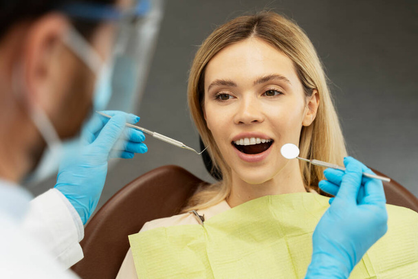 Professzionális fogorvos ellenőrzi a fogak minőségét. Fiatal, gyönyörű, mosolygós nő, páciens, aki a modern fogászati klinikán ül. Fogkezelés, egészségügyi koncepció  - Fotó, kép