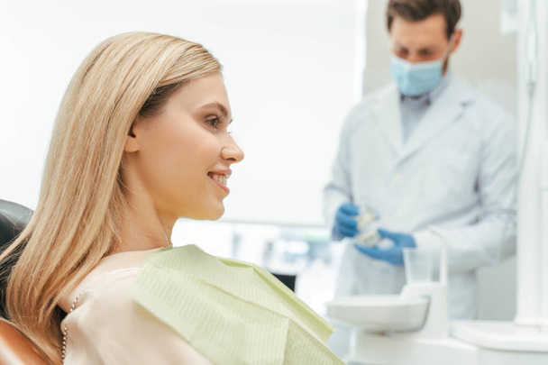 Młoda, piękna, uśmiechnięta kobieta, pacjent siedzący w nowoczesnej klinice stomatologicznej z dentystą w tle. Leczenie stomatologiczne, koncepcja opieki zdrowotnej  - Zdjęcie, obraz
