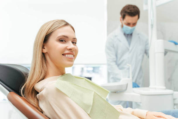 Jonge mooie glimlachende vrouw, patiënt zittend in moderne tandheelkundige kliniek met tandarts op de achtergrond. Tandheelkundige behandeling, gezondheidszorgconcept  - Foto, afbeelding