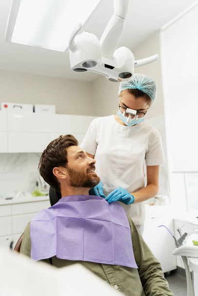 Portret atrakcyjnego uśmiechniętego pacjenta siedzącego na krześle dentystycznym rozmawiającego z dentystą. Atrakcyjna lekarka w lornetce zakładająca serwetkę dentystyczną. Leczenie, koncepcja opieki dentystycznej - Zdjęcie, obraz