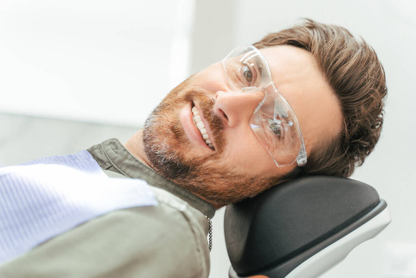 Όμορφος χαμογελαστός γενειοφόρος άντρας με προστατευτικά γυαλιά κάθεται στην οδοντιατρική καρέκλα, κοιτάζοντας την κάμερα. Ασθενής επισκέπτεται οδοντιατρική κλινική. Έννοια της οδοντιατρικής περίθαλψης - Φωτογραφία, εικόνα