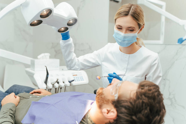 Erkek dişlerini kontrol ederken lambaya dönüşen profesyonel dişçi. Modern diş kliniğinde ayna ekipmanı tutan güzel bir bayan doktor. Diş tedavisi, bakım konsepti  - Fotoğraf, Görsel