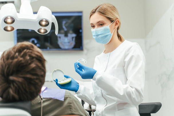 Dost canlısı kadın dişçi portresi, koruyucu yüz maskesi takan ortodontist hastaya hizalama gösteriyor. Diş sağlığı, diş sağlığı, diş koruma konsepti  - Fotoğraf, Görsel