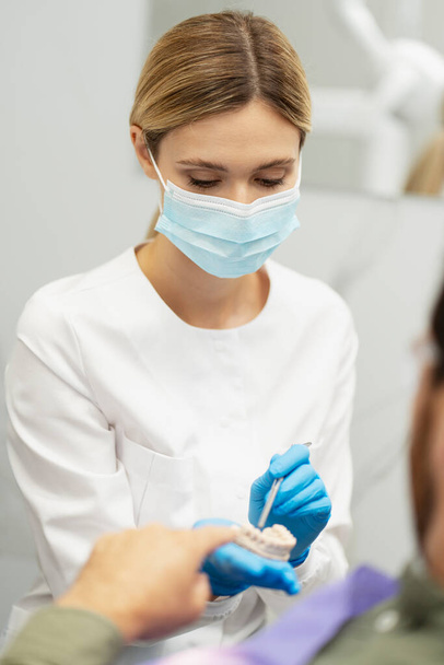 Mujer rubia dentista en guantes quirúrgicos azules, sosteniendo el modelo de mandíbula humana, mientras explica el tratamiento dental al paciente masculino en la clínica de odontología  - Foto, Imagen