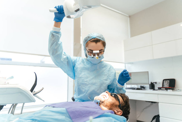 Kafasında mikroskop olan kadın cerrah erkek hasta için diş ameliyatı yapıyor. Kliniğe diş implantı ya da diş çıkarma kurulumu. Ortodonti ameliyatı sırasında genel anestezi.  - Fotoğraf, Görsel
