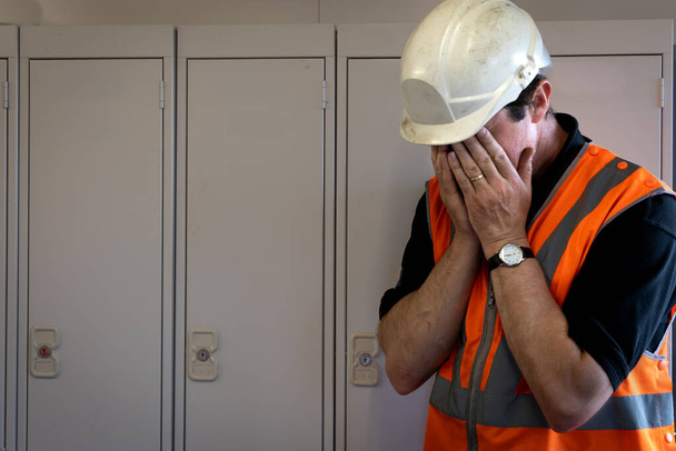 Een mannelijke werknemer met zijn hoofd in zijn handen in wanhoop worstelend om te gaan op het werk in een industriële of bouwomgeving in een concept van geestelijke gezondheid - Foto, afbeelding