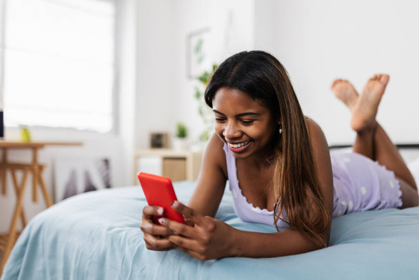jong latijn tiener student meisje met behulp van smartphone terwijl ontspannen op bed thuis. Millennial vrouw chatten op mobiele telefoon via social media app - Foto, afbeelding