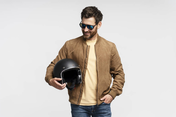 Handsome smiling bearded man, biker holding motorcycle helmet isolated on gray background. Fashion model wearing brown leather jacket, stylish sunglasses posing for pictures  - Φωτογραφία, εικόνα