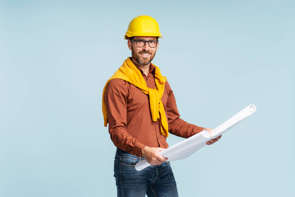 Porträt eines gut aussehenden lächelnden Architekten oder Ingenieurs mit gelbem Hut, der eine Architekturzeichnung isoliert auf blauem Hintergrund hält  - Foto, Bild
