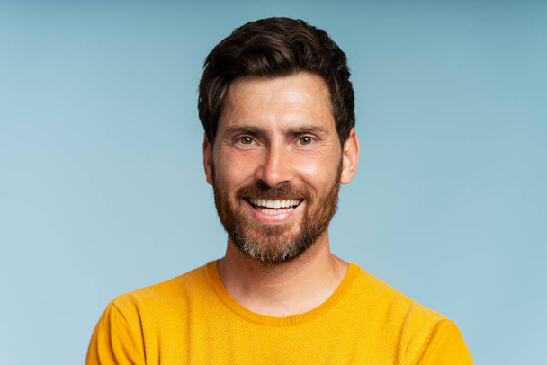 Портрет улыбающегося бородатого мужчины средних лет, смотрящего на камеру на голубом фоне. Уверенный в себе современный парень со стильными волосами позирует для фотографий - Фото, изображение