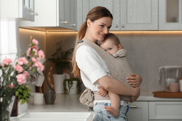 Mother holding her child in sling (baby carrier) in kitchen - Fotó, kép