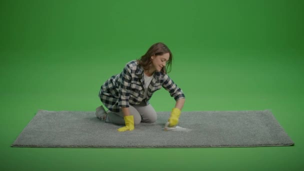 Een glimlachende jonge vrouw huisvrouw in geel beschermende rubberhandschoenen reinigt het tapijt met een borstel. Milieuvriendelijke reiniging. Onderhoud en reparatie van doe-het-zelfapparatuur. - Video