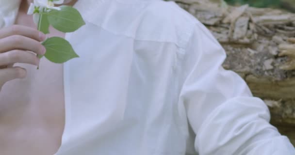 Nuori mies vaaleissa valkoisissa vaatteissa valkoinen kukka kädessään istuu vihreässä kukkivassa puutarhassa. Hidas liike - Materiaali, video