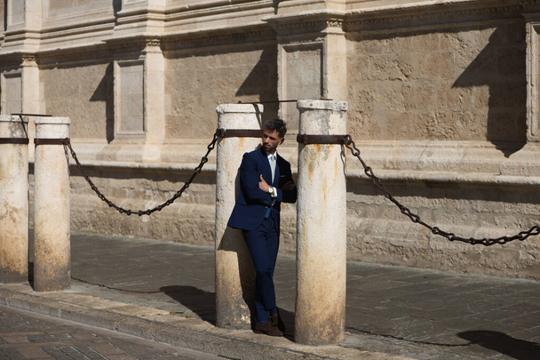 Hezký mladík s plnovousem, v modrém obleku a kravatě, zkříží paže opřené o sloup katedrály ve Španělsku. Ten muž je manažer, který je ve městě na služební cestě. - Fotografie, Obrázek