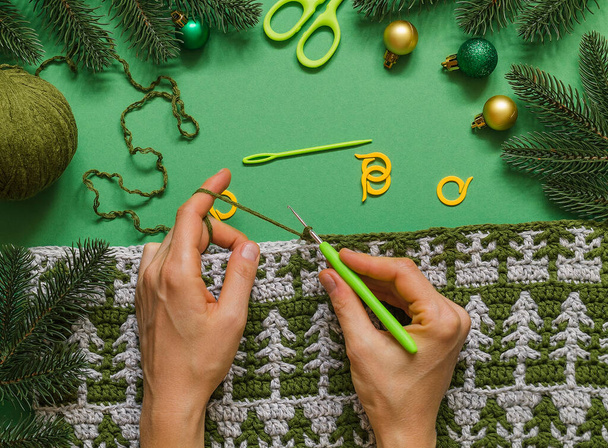 Woman's hands hold crochet hook and crochet fabric with Christmas tree ornament on a green background. - Photo, Image