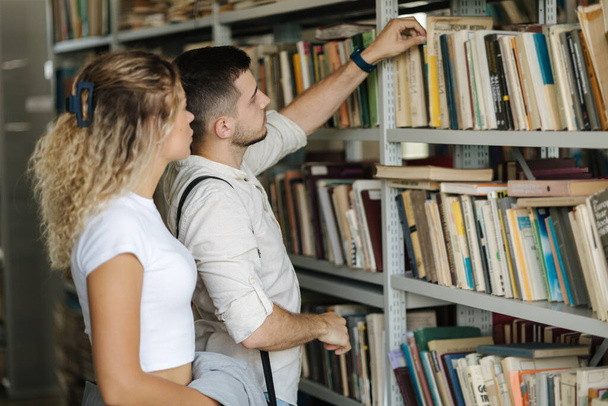 Man take book from book shelf to show his girlfriend. Classmates in college library . High quality photo - Photo, Image