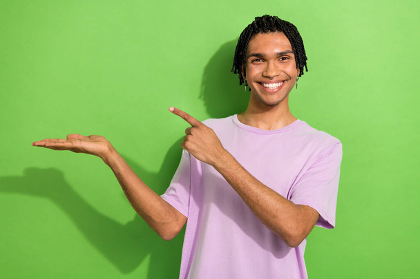 Photo of cheerful positive man dressed purple t-shirt holding arm pointing empty space isolated green color background. - Foto, afbeelding
